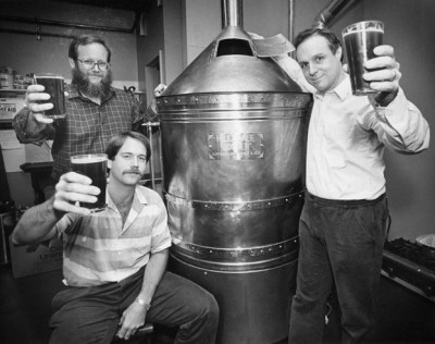 Three men holding full beers in toast around a copper brewing kettle